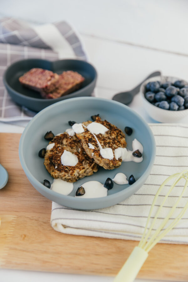 Blue silicone suction plate with banana pancakes on and yoghurt and blueberries.