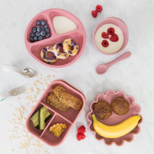 Scrummy Tummies divider plate, bowl and spoon, lunch box and flower plate all displayed with food inside and surrounding the products for display purposes.